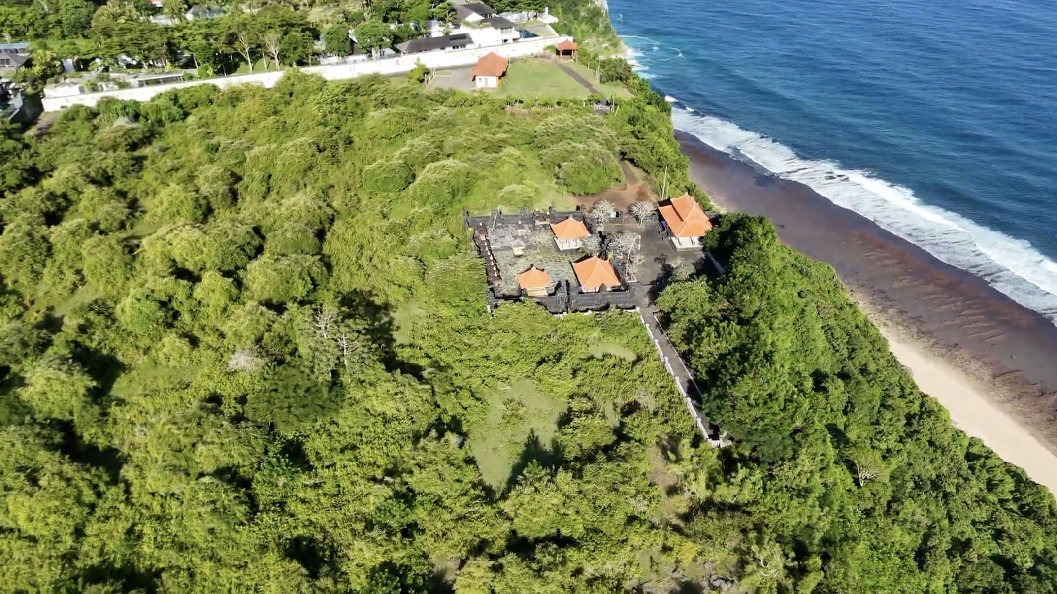 Aerial view along Uluwatu's forested limestone cliffs above the Indian Ocean and a long sandy beach