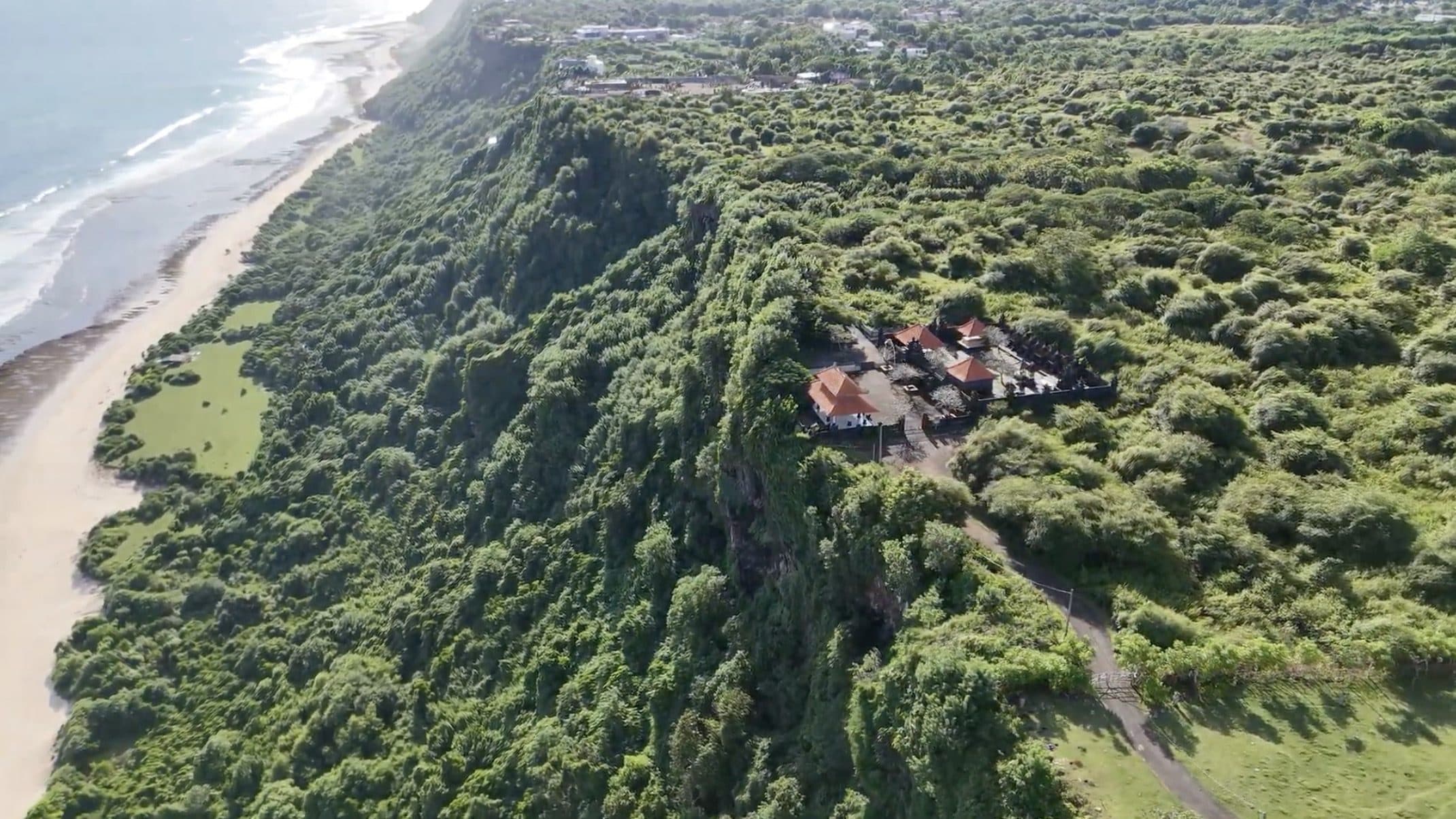 Aerial view of the south Bali coast with surf breaking on a wide beach below green cliffs